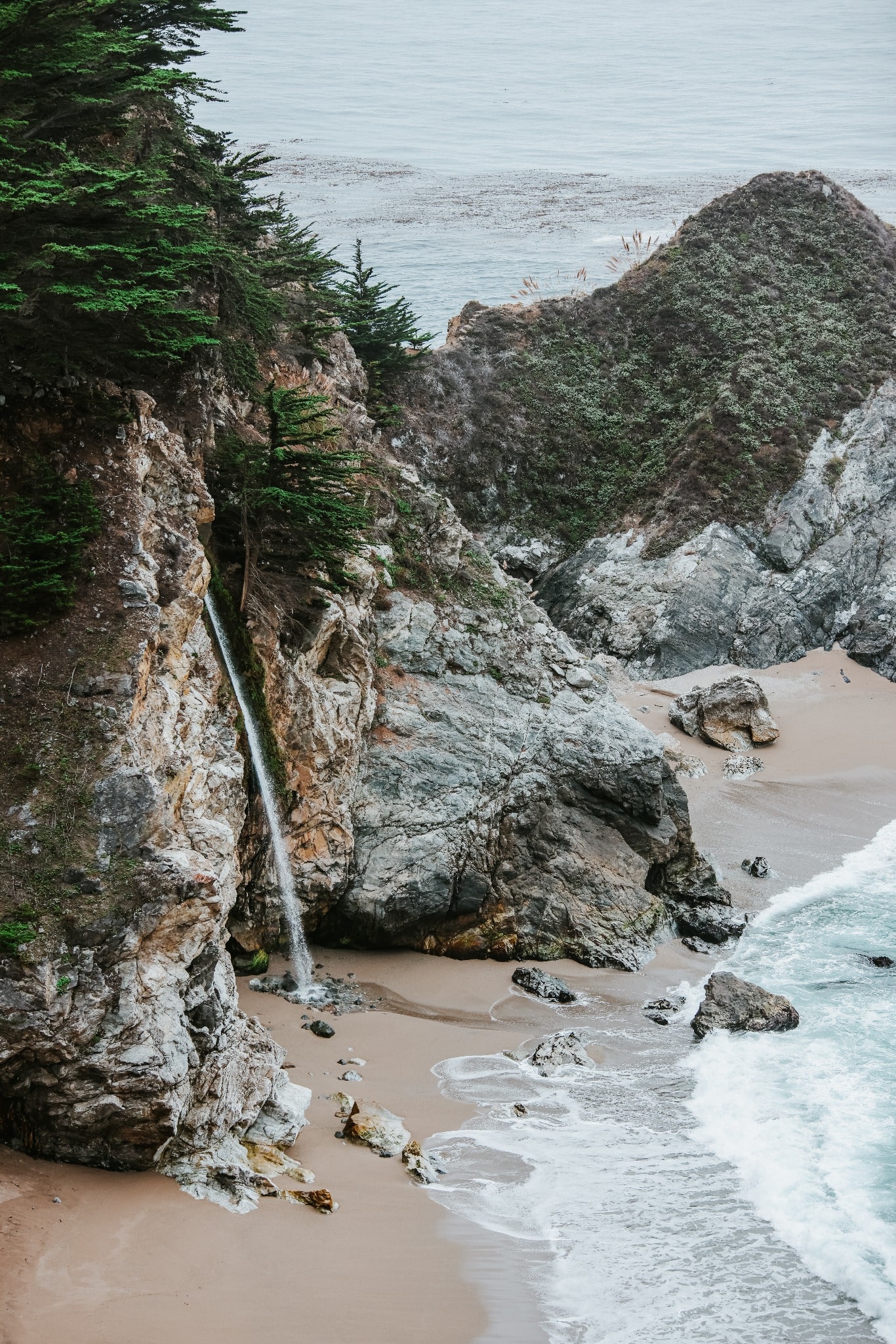 A waterfall drops from a forested coastal cliff onto a small beach beside sea-carved rock and the Pacific Ocean.