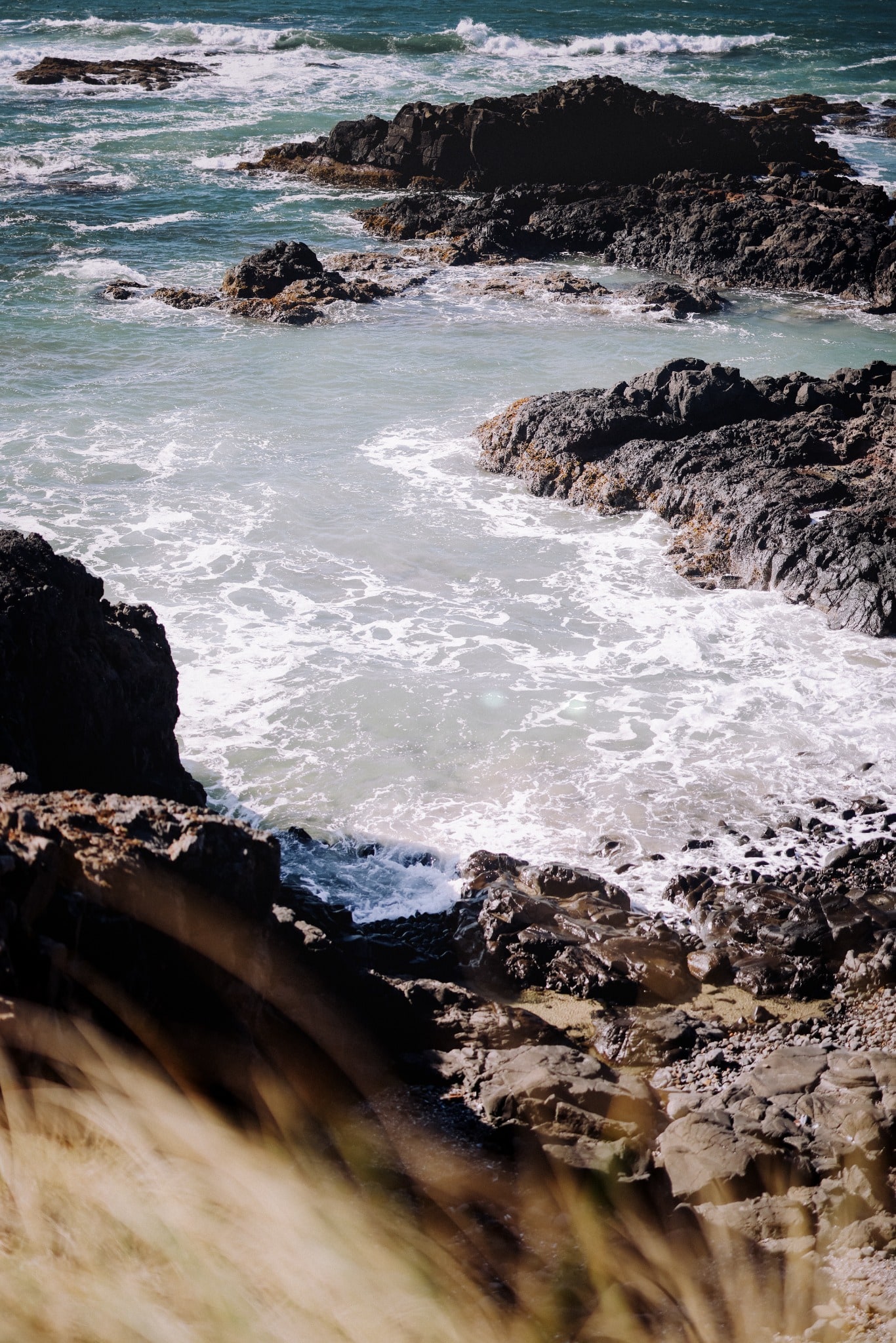 White surf courses between dark coastal rocks while teal waves break farther offshore.