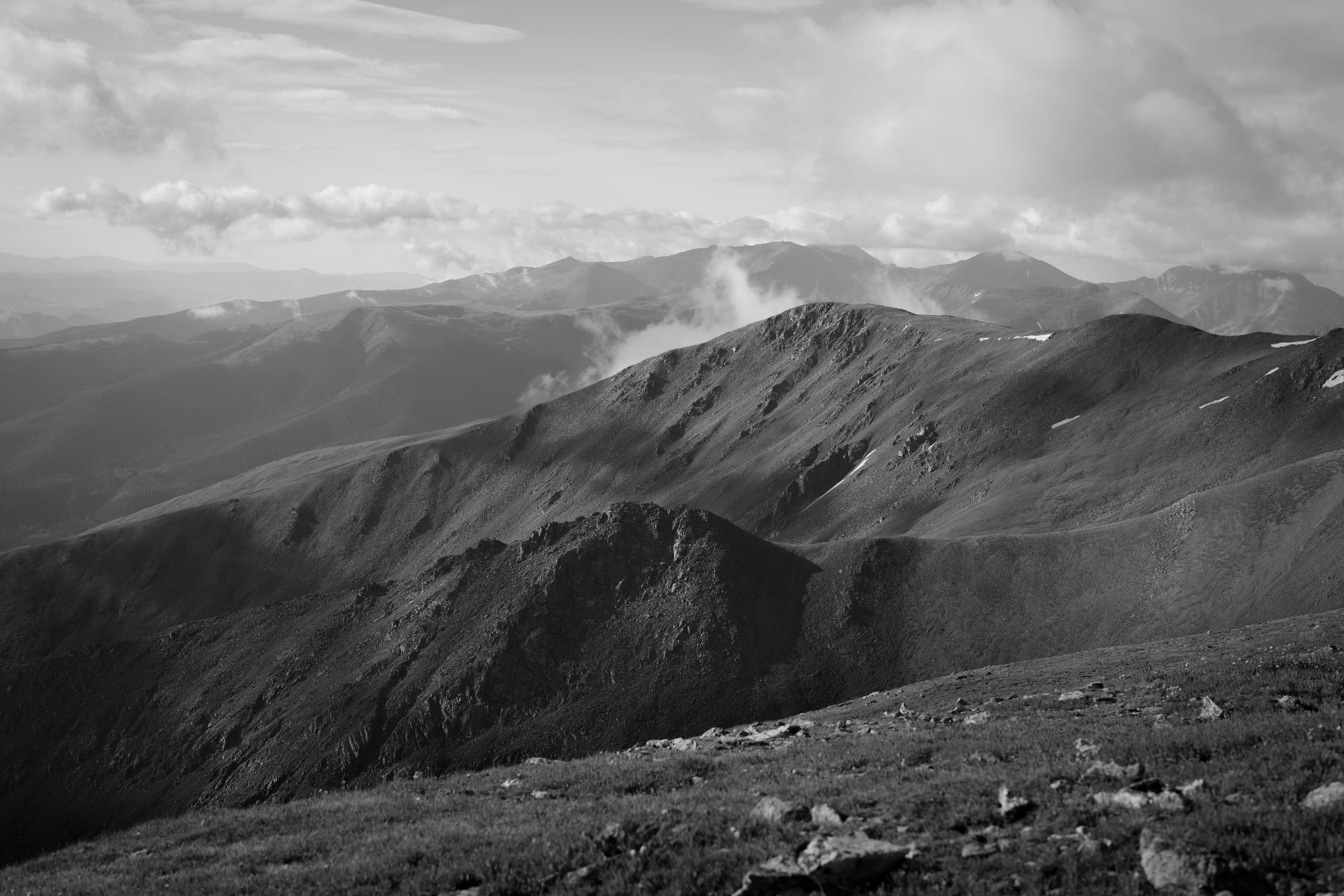 A black-and-white view across a high rocky ridge toward layered mountain ranges beneath bands of cloud.