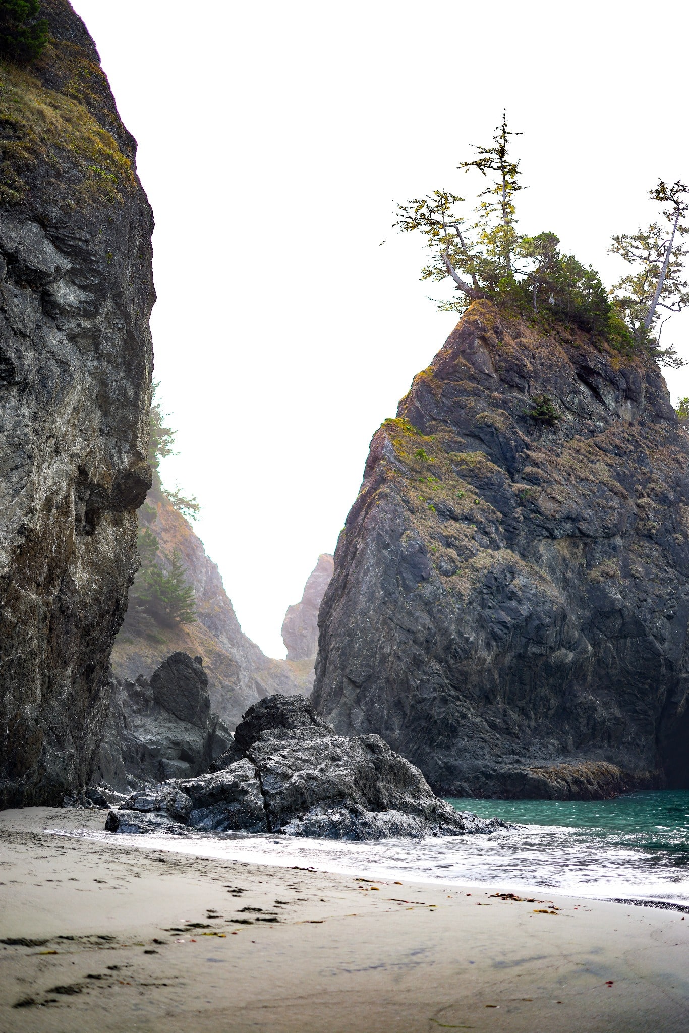 A narrow coastal passage opens between dark rock walls toward a pointed sea stack and pale overcast sky.