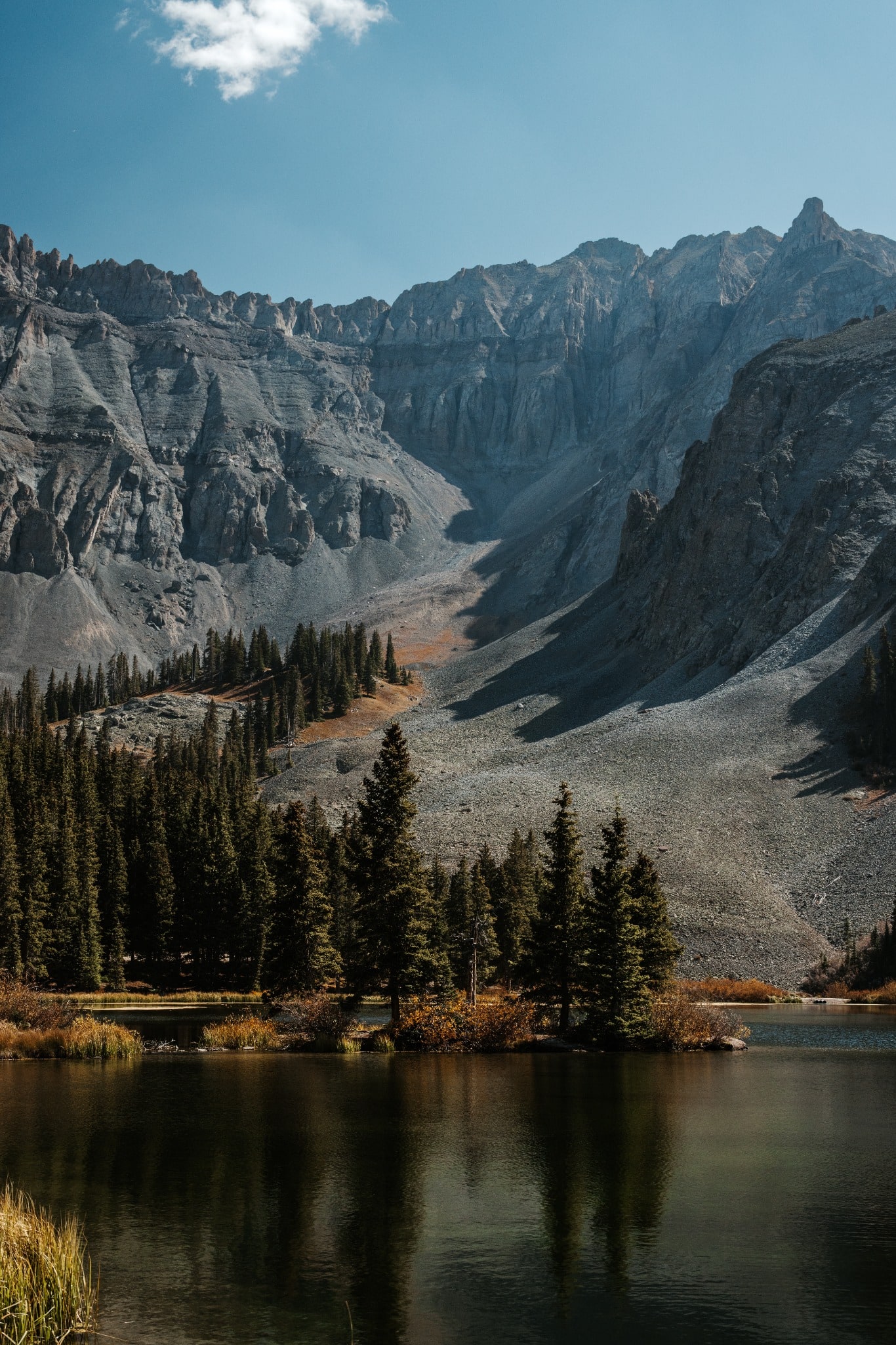 A calm alpine lake reflects a rugged mountain cirque and a stand of dark evergreen trees beneath a blue sky.