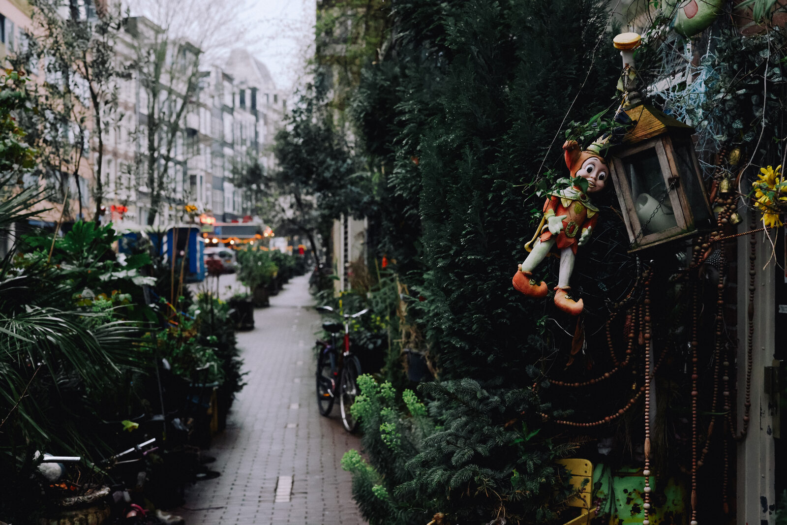 A narrow Amsterdam street lined with potted plants and a parked bicycle, a whimsical puppet ornament hanging on the right.