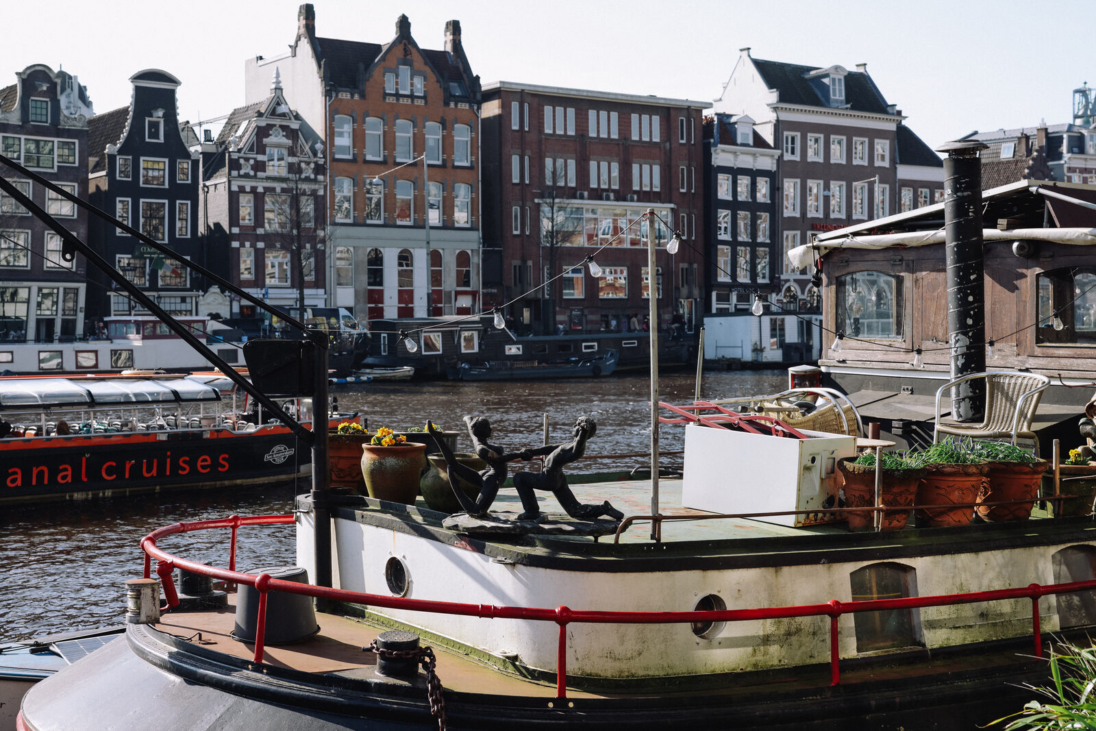 A canal houseboat with potted flowers in the foreground, gabled Amsterdam townhouses and a passing tour boat across the water.