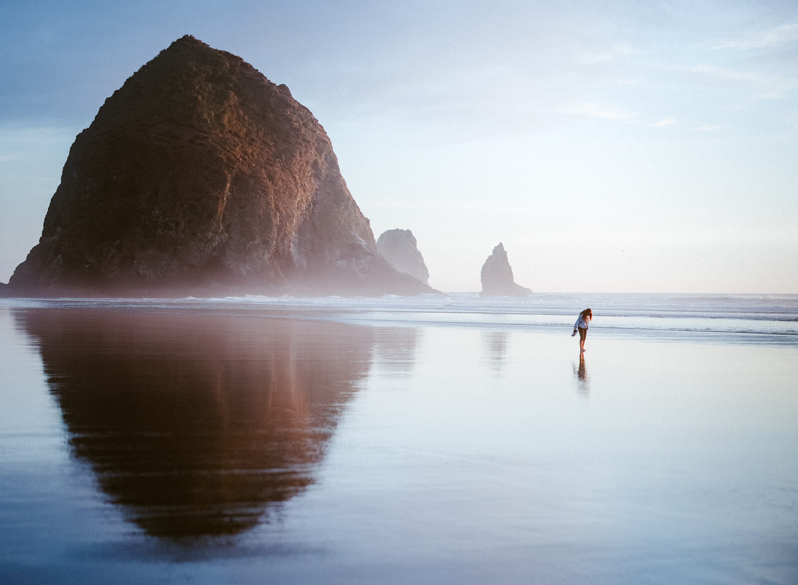 A lone figure walks across wet, reflective sand toward Haystack Rock and smaller sea stacks, soft blue morning light.