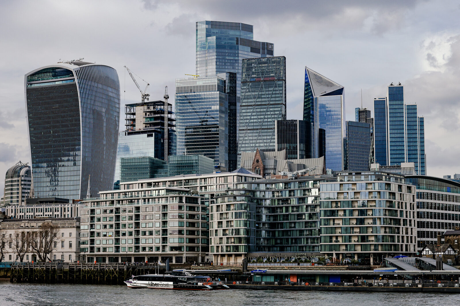 Glass skyscrapers of the City of London seen across the Thames, with cranes and a residential riverside development in the foreground.