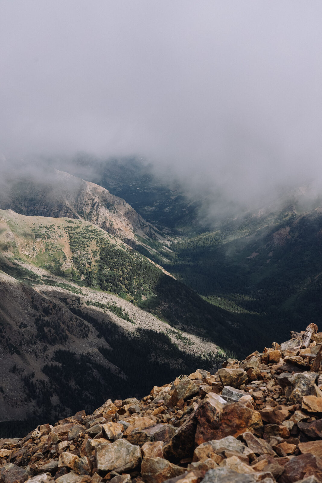 A misty, fog-wreathed mountain valley seen from a rocky summit, layered ridgelines fading into cloud.