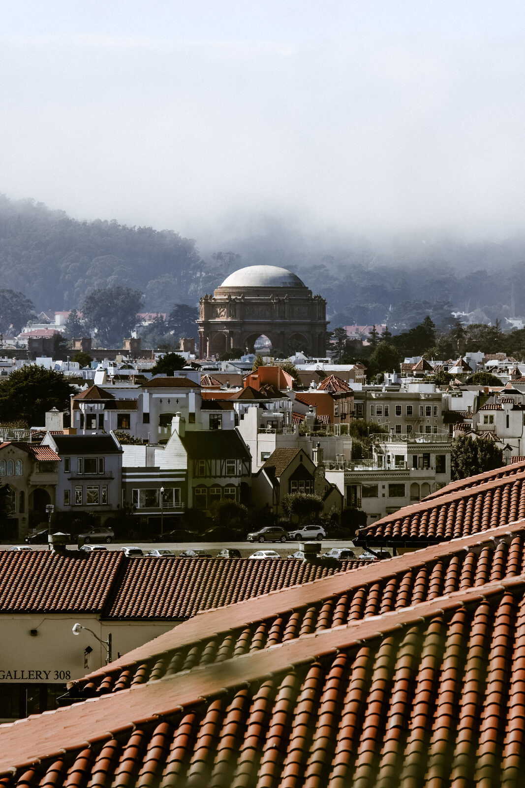 The domed rotunda of the Palace of Fine Arts rises above fog-covered hills and rooftops in a San Francisco neighborhood.